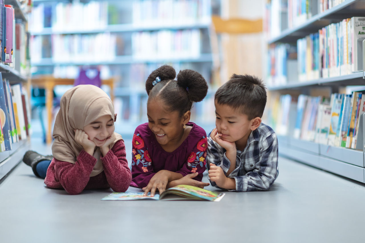 Books to Celebrate Black History Month Three children of diverse cultural background lying on the floor of a library reading a book together