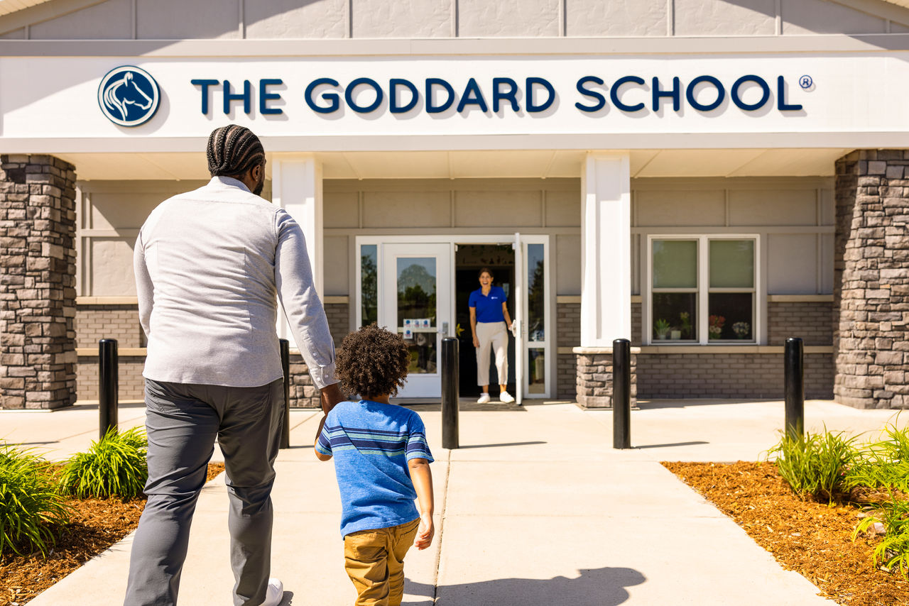 Why Families Choose The Goddard School A father and son holding hands walking up to a Goddard School entryway where a teacher is waiting to greet them.